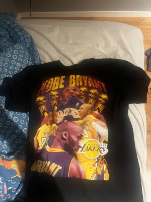Svart Kobe Bryant Lakers t-shirt - Cool svart t-shirt med stort tryck av Kobe Bryant och Los Angeles Lakers på både fram- och baksidan. Flera motiv av Kobe i olika Lakers-tröjor och med basketbollar samt text i gult och lila. Perfekt för basketfans som vill sticka ut.