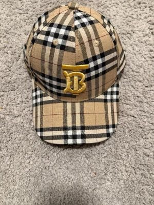 Burberry keps med klassiskt rutmönster - Snygg keps från Burberry i deras ikoniska beige, svarta och röda rutmönster. Framtill sitter ett broderat gult 'TB'-monogram. Justerbar baktill och tillverkad i bomull. Perfekt accessoar för att lyfta din streetwear-look.