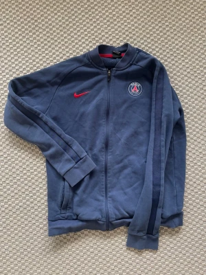 Blå PSG träningsjacka från Nike - Mörkblå träningsjacka från Nike med PSG-logga på bröstet och röd Nike-swoosh. Jackan har hel dragkedja, ribbade muddar och mörkblå ränder längs ärmarna. Perfekt för fotbollsträning eller som sportig vardagsstil.