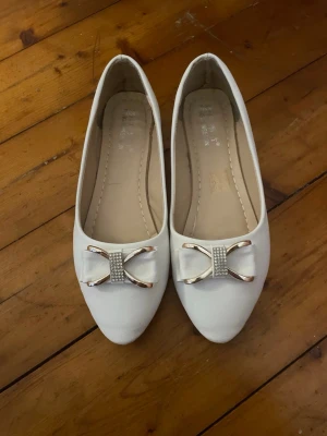 Vita ballerinaskor med rosett - Snygga vita ballerinaskor med spetsig tå och dekorativ rosett framtill i silver och små glittrande stenar. Skorna har låg klack och är tillverkade i ett slätt material som ger en stilren look. Aldrig använda! 