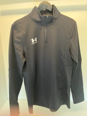 Svart träningströja från Under Armour - Snygg svart långärmad träningströja från Under Armour med halv dragkedja och hög krage. Perfekt för löpning eller gym. Tillverkad i ett mjukt och stretchigt funktionsmaterial som andas. Diskret logga på bröstet.