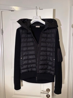 Moncler Cardigan storlek Large - Snygg svart Moncler Cardigan med stickade ärmar och huva. Jackan är väldigt sparsam använd och är i mycket bra skick. Finns inga defekter eller skador.                                                                 Kostar 12 045 kr i butik. 
