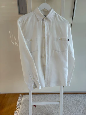 Vit linneskjorta från Lexington - Klassisk vit skjorta från Lexington i svalt linne. Skjortan har lång ärm, två bröstfickor och knappar framtill. Perfekt för dig som gillar en clean och stilren look. Fint skick, dock en liteskreva på ärm. Se bild. Kan lätt lagas och syns inte om man viker upp ärmen.