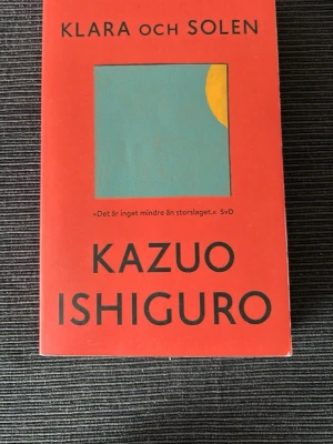 Klara och solen av Kazuo Ishiguro - En skönlitterär roman med röd pocketomslag, skriven av Kazuo Ishiguro. Boken utforskar teman om kärlek och mänsklighet genom roboten Klara. Omslaget har en minimalistisk design med en blå kvadrat och gul cirkel. Perfekt för dig som gillar tankeväckande berättelser.