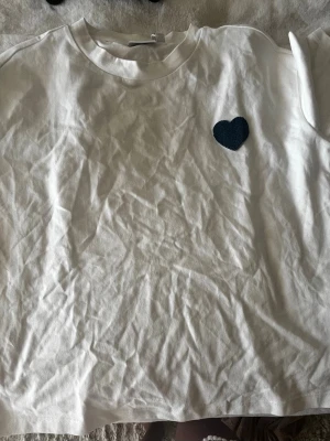 Vit t-shirt med hjärtdetalj från Vila - Vit t-shirt från Vila med en mörkblå hjärtapplikation på bröstet. Klassisk rund halsringning och korta ärmar. Tillverkad i mjuk bomull för en skön känsla. Perfekt för en enkel och stilren look. Den ska var lite oversized skulle säga att den var i storlek M men är jättesnygg. Funkar på person som har storlek S också.💞