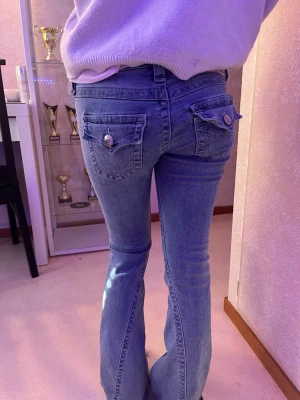 True Religion bootcut jeans ljusblå - Säljer ett par ljusblå bootcut jeans från True Religion med ikoniska fickor bak och snygga detaljer i sömmarna. Jeansen har knappar med logga och klassisk patch bak i midjan. Perfekta för dig som gillar en avslappnad men trendig stil.