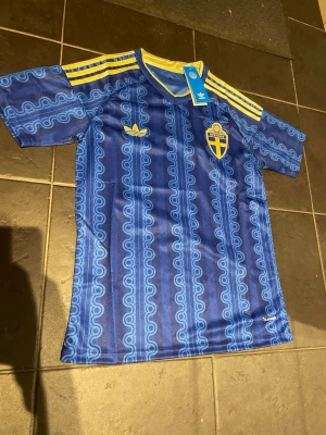 Blå Sverige fotbollströja Adidas - Snygg blå fotbollströja från Adidas med Sveriges landslagsemblem på bröstet och gula detaljer. Tröjan har ett vågigt mönster i olika blå nyanser, gula Adidas-ränder på axlarna och är tillverkad i ett lätt, ventilerande material.