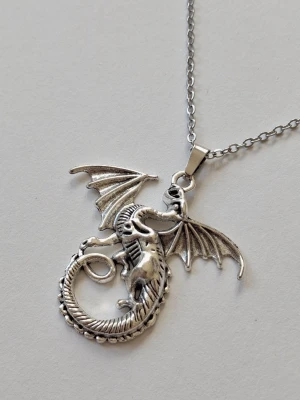 Halsband med drakberlock i silverfärg - Unikt halsband (ankarlänk) med en detaljerad drakberlock i silverfärgad metall. Draken har utbredda vingar och en böjd kropp, vilket ger ett coolt och fantasyinspirerat uttryck. Perfekt accessoar för dig som gillar mytologi och fantasy. Materialet rostfritt stål. 