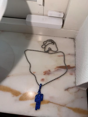 Halsband med blå glittrig blohsh - Snyggt halsband med en silverfärgad kedja och en blå glittrig berlock i form av en stiliserad blohsh. Inspireras av billie eilish !! Halsbandet är 55 cm långt, och inte använt ❤️ 