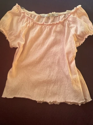 Ljusrosa offshoulder blus - Söt ljusrosa blus med offshoulder-modell och korta ärmar. Toppens material är lätt och har en skrynklig struktur, vilket ger en avslappnad och trendig känsla. Perfekt att matcha med jeans eller kjol för en fräsch look.