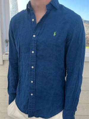 Blå linneskjorta från Ralph Lauren - Säljer en snygg blå skjorta från Ralph Lauren i linne med klassisk button-down krage och vita knappar. Skjortan har lång ärm och den ikoniska gröna loggan broderad på bröstet. Perfekt för en stilren och avslappnad look.