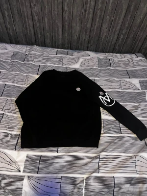 Svart Moncler sweatshirt med logga - Säljer en svart sweatshirt från Moncler med rund hals och vita logodetaljer på bröstet och ena ärmen. Tröjan har en avslappnad passform och är tillverkad i mjukt material, perfekt för en trendig och bekväm stil.