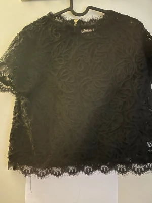 Svart spetsblus med broderade detaljer - Säljer en svart blus med korta ärmar i genombruten spets och broderade mönster. Blusen har rund halsringning och fina spetskanter nertill. Perfekt för dig som gillar unika och detaljerade toppar.