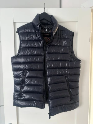Svart dunväst från Save The Duck - Snygg svart dunväst från Save The Duck med glansig finish och horisontella sömmar. Västen är tillverkad av 100% återvunnet material och har två dragkedjeförsedda fickor. Perfekt för lager-på-lager och ett medvetet val för miljön. Använd max 5 gånger. 