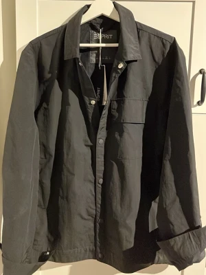 Svart overshirt från Esprit - Stilren svart overshirt från Esprit med klassisk krage, tryckknappar och bröstficka. Overshirten har en rak passform och är tillverkad i ett lätt material som passar perfekt för lager-på-lager. Snygg och enkel att matcha med olika outfits.