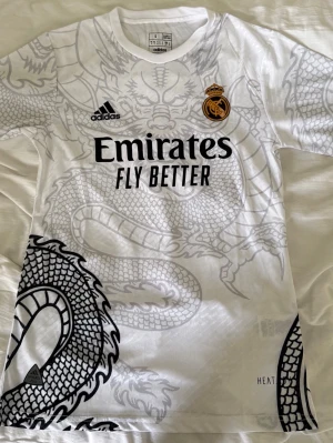 Real Madrid x Adidas draktröja - Snygg vit Real Madrid-tröja från Adidas med stort drakmönster i grått och svart över hela plagget. Tröjan har klubbmärke, Adidas-logga och Emirates Fly Better-tryck på bröstet. Tillverkad i lätt och ventilerande material, perfekt för fotboll eller träning.