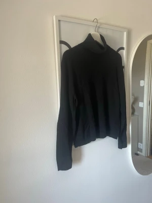 Svart polotröja i stickat material - En svart stickad polotröja med lång ärm och hög krage. Tröjan har en klassisk och enkel design som passar till många olika stilar. Perfekt för lager-på-lager och snygg till både jeans och kjol.