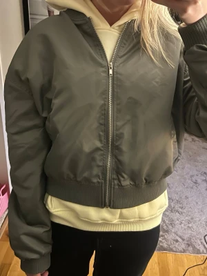 Olivgrön bomberjacka med dragkedja - Snygg olivgrön bomberjacka med klassisk passform och ribbade muddar. Jackan har två fickor framtill och stängs med dragkedja. Perfekt för dig som vill ha en stilren och enkel look. Materialet är glansigt och ger en modern känsla.