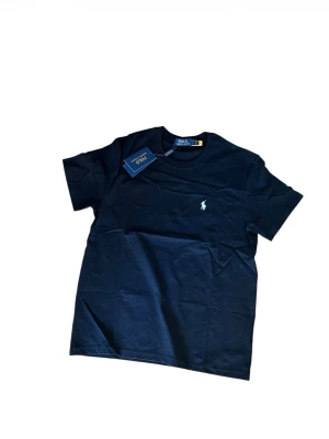 Mörkblå t-shirt från Polo Ralph Lauren - Snygg svart t-shirt från Polo Ralph Lauren med klassisk passform och rund hals. T-shirten har en liten vit broderad logga på bröstet och är tillverkad i mjuk bomull. Perfekt för en clean och stilren look.