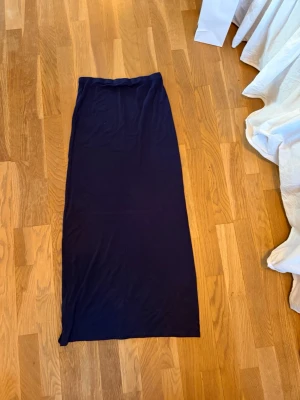 Mörkblå långkjol från Nelly - En stilren mörkblå långkjol från Nelly. Kjolen har en rak och enkel design med resår i midjan och är tillverkad i ett mjukt och lätt material som känns skönt mot huden. Perfekt för dig som gillar minimalistisk stil.