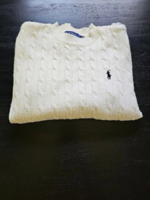 Vit kabelstickad tröja Polo Ralph Lauren - Kabelstickad vit tröja från Polo Ralph Lauren med rund halsringning och klassisk broderad logga på bröstet. Tröjan har ribbstickade muddar och nederkant, perfekt för en stilren och tidlös look.
