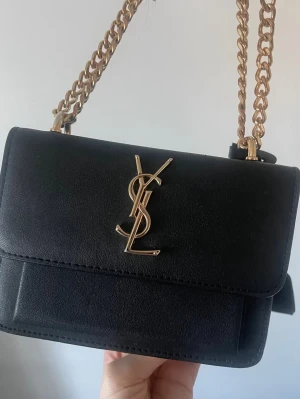 Saint Laurent sunset bag - Otroligt fin i läder, svart med guldiga detaljer. Går att använda på många olika sätt, med lång kedja, över axeln, som clutch eller som jag gjort på bild, med en kortare kedja på handen. Tyvärr inget kvitto på väskan då den är köpt vintage, men dustbag tillkommer. Fint skick, med några få slitage men endast synligt om man tittar noga. Inget som syns mycket alls.