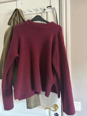Vinröd stickad tröja från Kappahl - Mysig vinröd stickad tröja från Kappahl med avslappnad passform, ribbade muddar och slitsar i sidorna. Perfekt för lager-på-lager och enkel att styla till jeans eller kjol. Rund halsringning och mjukt material.