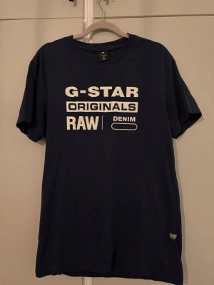 Blå G-Star RAW t-shirt med tryck - Snygg marinblå t-shirt från G-Star RAW med vit logotyp och texttryck framtill. Klassisk rund halsringning och korta ärmar. 
