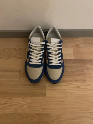 Philippe Model blåvita sneakers - Snygga sneakers från Philippe Model i blått och vitt med beige detaljer. Ovandelen är i en mix av mocka och mesh, vilket ger en sportig och fräsch look. Klassisk låg modell med vita snören och vit sula. Perfekta för dig som gillar stilrena och trendiga skor.