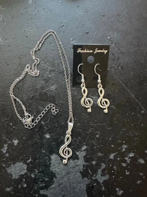 Smyckesset med silverfärgade noter - Snyggt smyckesset med halsband och örhängen i silverfärgad metall. Både halsbandet och örhängena har hängen formade som G-klavar, perfekt för dig som älskar musik. Kedjan är tunn och justerbar. Passar till att lyfta vilken outfit som helst.