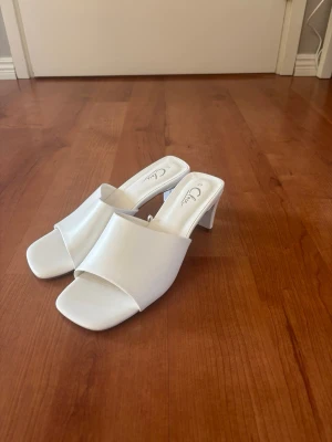 Vita slip-in sandaler med klack - Stilrena vita sandaler från Clou med öppen tå och fyrkantig front. Slip-in-modell med bred rem och medelhög klack. Tillverkade i ett slätt material som ger en clean look. Perfekta för dig som gillar minimalistisk och trendig stil. Använda en gång.