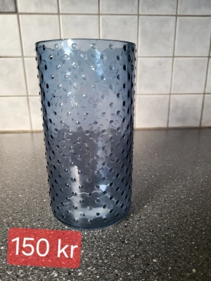 Blå vas med prickig struktur - Säljer en hög blå vas med prickig struktur på utsidan. Glaset har en cylindrisk form och är tillverkat av transparent blått glasmaterial. 
