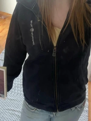 Svart hoodie från Sail Racing - Snygg svart hoodie från Sail Racing med dragkedja framtill och logga på bröstet. Hoodien har huva med dragsko och en bröstficka med dragkedja. Tillverkad i mjukt material som passar perfekt till en avslappnad stil.