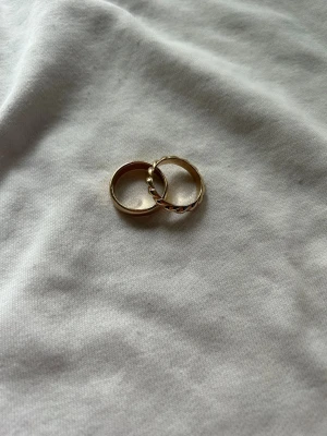 Två guldfärgade ringar - Helt oanvända ringar från Ur&Penn. Två stilrena guldfärgade ringar. Den ena ringen har en slät, klassisk design medan den andra har en tvinnad form med små pärldetaljer längs kanten. Perfekta att bära tillsammans eller var för sig för en trendig look.