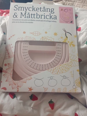 Smycketång och mättbricka rosa - Säljer en rosa smycketång och mättbricka i plast, perfekt för att hålla koll på pärlor och tillbehör när du gör egna smycken. Brickan har tydliga måttmarkeringar och flera spår för att sortera pärlor. Kommer i originalförpackning med dekorativ design.