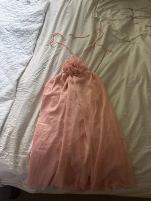 Ljusrosa klänning - En superfin ljusrosa klänning med tunna band som knyts ihop bakom nacken. Klänningen har ett luftigt fall och är tillverkad i ett tunt, mjukt tyg som ger en elegant känsla. Perfekt för sommarens speciella tillfällen.