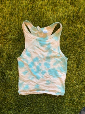 Turkos och vit tie-dye linnetopp - Snygg linnetopp i vitt med egengjorda turkosa batikmönster. Modellen är ärmlös med brottarrygg och croppad passform. Väldigt liten.