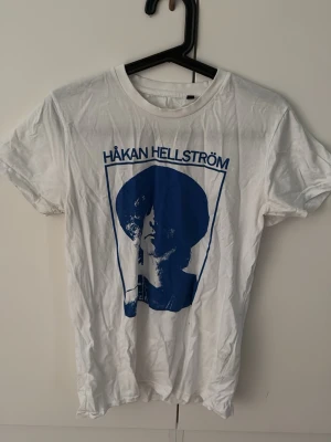 Vit Håkan Hellström t-shirt med tryck - Säljer en vit t-shirt med blått tryck av Håkan Hellström på framsidan. T-shirten har rund hals och korta ärmar, perfekt för dig som vill visa din kärlek till svensk musik. Materialet är mjuk bomull och passformen är normal.