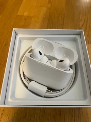 Apple AirPods Pro med laddningsetui - Säljer ett par vita  Pro andra generationen med trådlöst laddningsetui. Hörlurarna har en stilren och kompakt design med silikonproppar för bra passform. Medföljer originalförpackning, laddningskabel och instruktioner. Perfekta för musik, samtal och träning.
