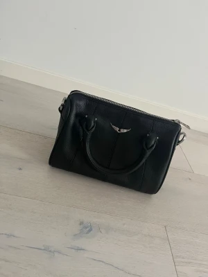 Svart handväska Zadig & Voltaire - Snygg svart handväska från Zadig & Voltaire i äkta läder. Väskan har två handtag, silverfärgade detaljer och en ikonisk logga med vingar framtill. Kommer med original dustbag. Perfekt för dig som gillar stilrena accessoarer med en rockig touch.