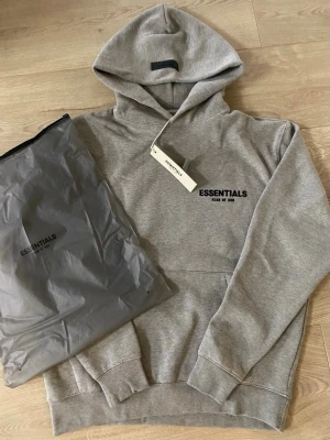 Grå Essentials Fear of God hoodie - Säljer en grå hoodie från Essentials Fear of God med svart logga på bröstet. Klassisk passform med huva och känguruficka framtill. Tillverkad i mjukt bomullsmaterial som är perfekt för en avslappnad och trendig look. Perfekt för dig som gillar streetwear.