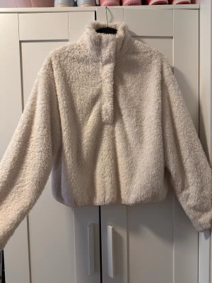 Gräddvit fluffig stickad tröja - Mysig och fluffig vit stickad tröja från H&M med hög krage och tryckknappar framtill. Tröjan har lång ärm och är tillverkad i ett mjukt, teddy-liknande material som håller dig varm under kyliga dagar. Perfekt för dig som gillar en avslappnad och trendig stil.