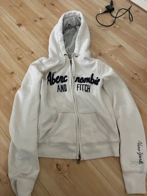 Abercrombie & Fitch - Vit hoodie med dragkedja från Abercrombie & Fitch. Stor huva, två fickor fram och broderad text på bröstet samt 'New York' på ärmen. Mjuk insida och ribbade muddar. Perfekt för en avslappnad och sportig stil. Finns en liten grön fläck på ena armen som syns på bilden men ska försöka få bort den i tvätten, behövde bara få ut annonsen snabbt då jag behöver pengarna till min hund🙈Skriv vid frågor!! Står att storleken är M men skulle säga att den passar mindre då jag brukar ha XS-S och den passar!