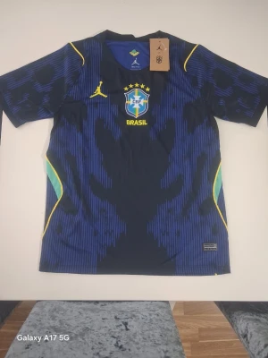 Blå Brasilien fotbollströja Jordan - Snygg blå Brasilien fotbollströja från Jordan med CBF-logga och fem stjärnor på bröstet. Tröjan har gula detaljer, Dri-FIT-material och ett unikt mönster i svart och blått. Perfekt för dig som älskar fotboll och vill sticka ut på planen.