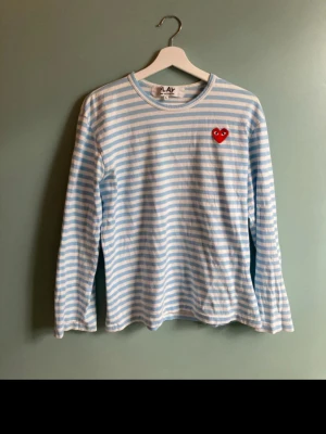 Randig tröja Comme des Garçons Play - Ljusblå och vit randig långärmad tröja från Comme des Garçons Play med det ikoniska röda hjärte-logotypen på bröstet. Tillverkad i mjuk bomull och har en avslappnad passform. Perfekt för dig som gillar streetwear och stilrena plagg. Sitter som M typ!