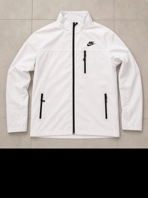 Vit vindjacka från Nike - Snygg vit vindjacka från Nike med svarta dragkedjor och logga på bröstet. Jackan har hög krage, tre fickor med dragkedja och är tillverkad i lätt, vindtätt material. Perfekt för dig som vill ha en clean och sportig look.