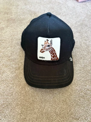 Svart truckerkeps med giraffpatch - Säljer en svart truckerkeps från The Farm med en broderad patch framtill som visar en giraff och texten 'HIGH'. Kepsen har mesh på baksidan för extra ventilation och justerbar snapback. Skärmen är brun undertill och kepsen har en cool streetstyle-vibe.