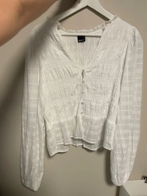 Vit blus med knappar från Gina Tricot - Säljer en söt vit blus från Gina Tricot med smockdetaljer och v-ringning. Den var tyvärr för stor, använd 1-2 gånger.