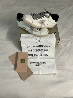 Golden Goose - Golden Goose sneakers i vitt läder med grå mockastjärna på sidan, grå skosnören och svart hälparti. Klassisk låg modell med slitna detaljer och beige sula. Kommer med originalpåse och box. Perfekta för dig som gillar streetstyle och exklusiva märken. Skorna är helt oanvända. 1: 1. Hade kunnat tänka mig att gå ner lite i pris vid snabb och smidig affär. Vid några funderingar är det bara att fråga. 