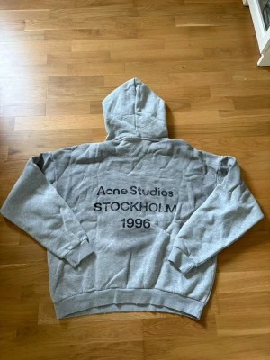 Grå hoodie Acne Studios Stockholm - Snygg grå hoodie från Acne Studios med tryck på ryggen: 'Acne Studios STOCKHOLM 1996'. Tillverkad i mjuk 100% bomull med borstad insida för extra komfort. Klassisk passform med huva och ribbade muddar.